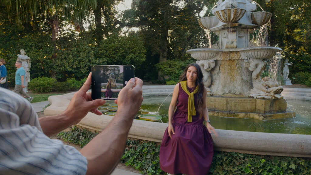 Someone holding iPhone Duo open to photograph a woman sitting by a fountain